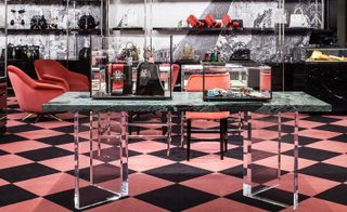 A grey marble table with clear plastic legs, glass display cases on top of it, a pink checkered floor below it and pink chairs around it.