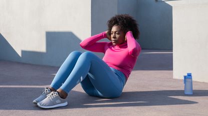 Woman performs sit-up exercise outside. She is wearing a pink long-sleeved top, blue leggings and grey trainers.