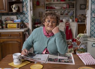 Mrs Brown's Boys Christmas specials 2024 star and creator Brendan O'Carroll in character as Mrs Brown, sitting in her kitchen and reading a newspaper, while smiling at the audience. 