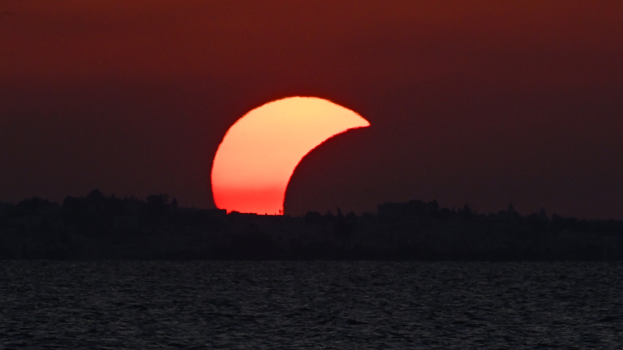 the sun appears as a crescent as it sets behind a tree line with a lake in the foreground
