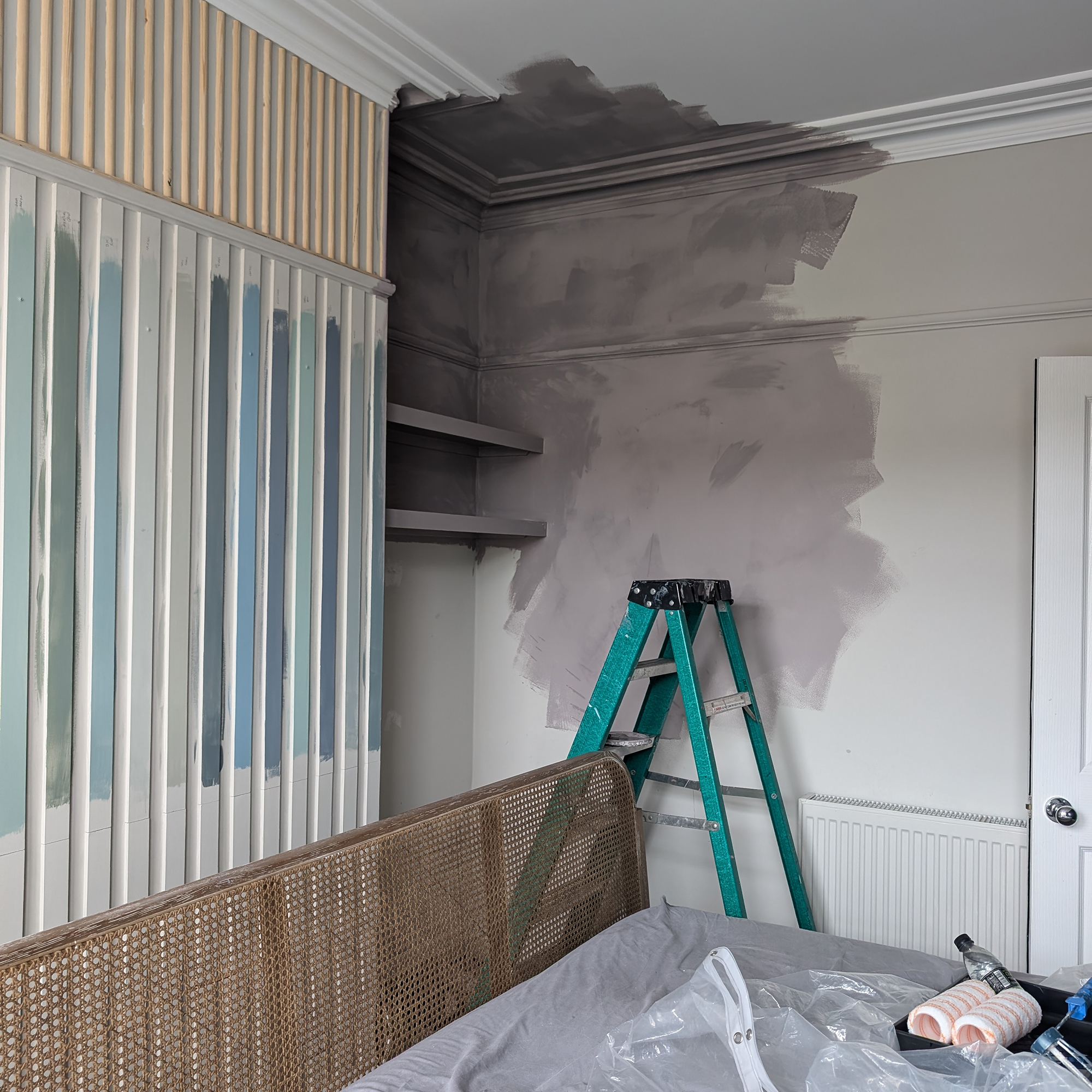 neutral bedroom with half painted wall and a green ladder standing in front of it and paint rollers on the bed