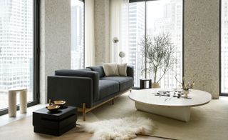 A photo of an apartment room with large floor to ceiling windows. Featured in the room is a navy sofa on the left, a round white coffee table in the middle and a navy blue side table next to the sofa. In the background is an indoor plant and lamp.