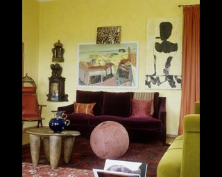 Small living room with warm yellow paint on walls and abstract art on the walls with purple and green velvet sofas and wooden accent furniture