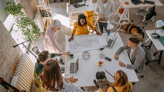 A small office team gather around a table to pore over architectural plans, representing digital transformation for small businesses. They are in a bright and modern office.