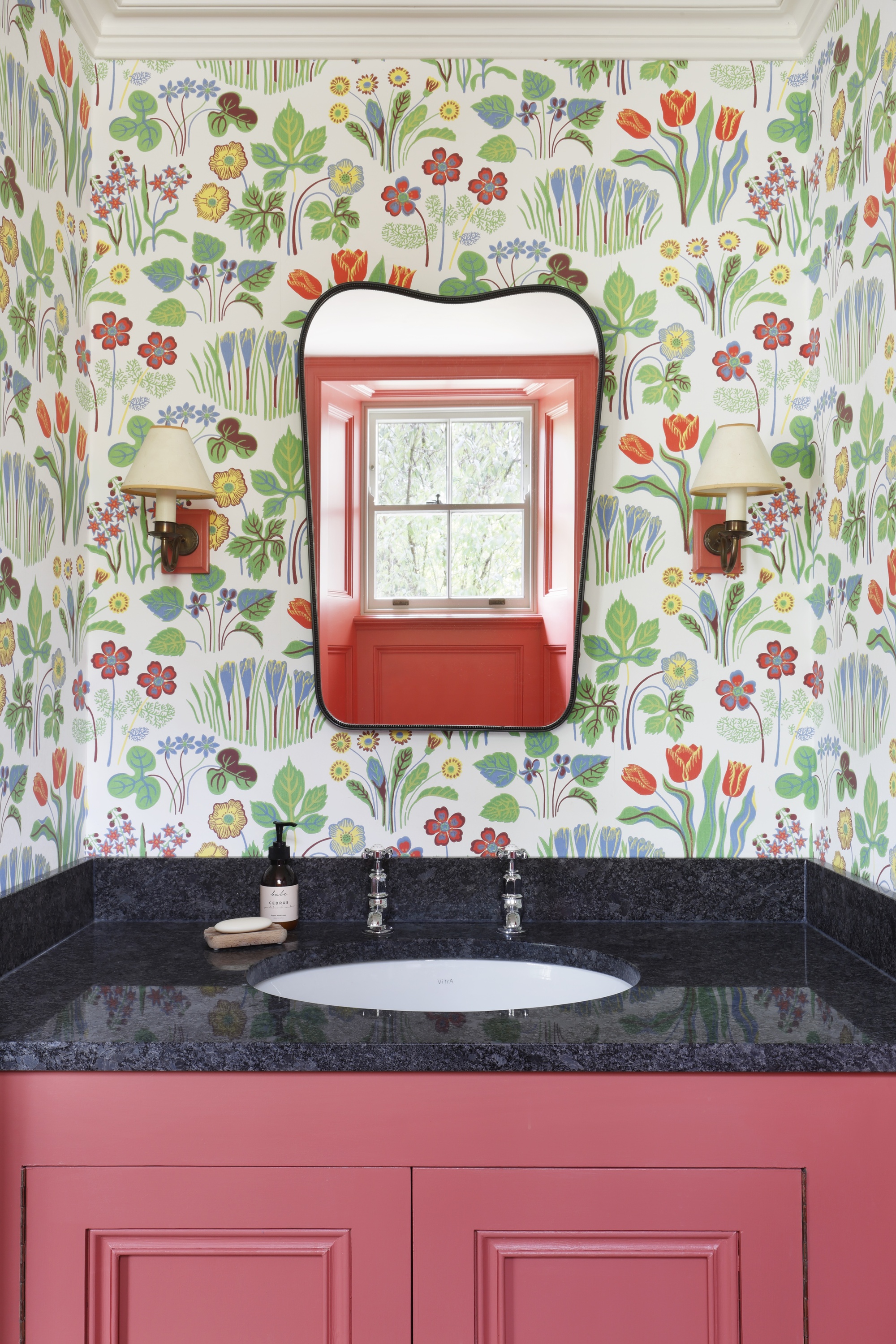maximalist bathroom with garden print wallpaper, red cabinetry, black counter, wall sconces, and black-rimmed mirror