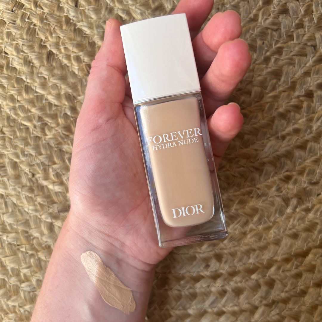Beauty Editor Fiona McKim's hand holding a tube of foundation, with a swatch of the foundation on her wrist, on a woven jute rug backdrop