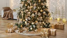 Image of the bottom half of Christmas tree covered in champagne and cream-colored ornaments, garland, and lights. There are brown paper packages around the tree and a white bench seat in the back.