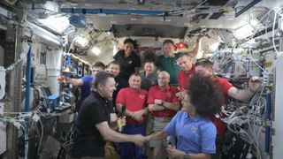 Change of command ceremony aboard the International Space Station, Sep. 22, 2024. A man in a black shirt shakes the hand of a woman in a blue shirt, inside a crowded room of wires and electronic gear. behind them, six people, four in red, one in grey, one in gree, one in black, and the last in blue.