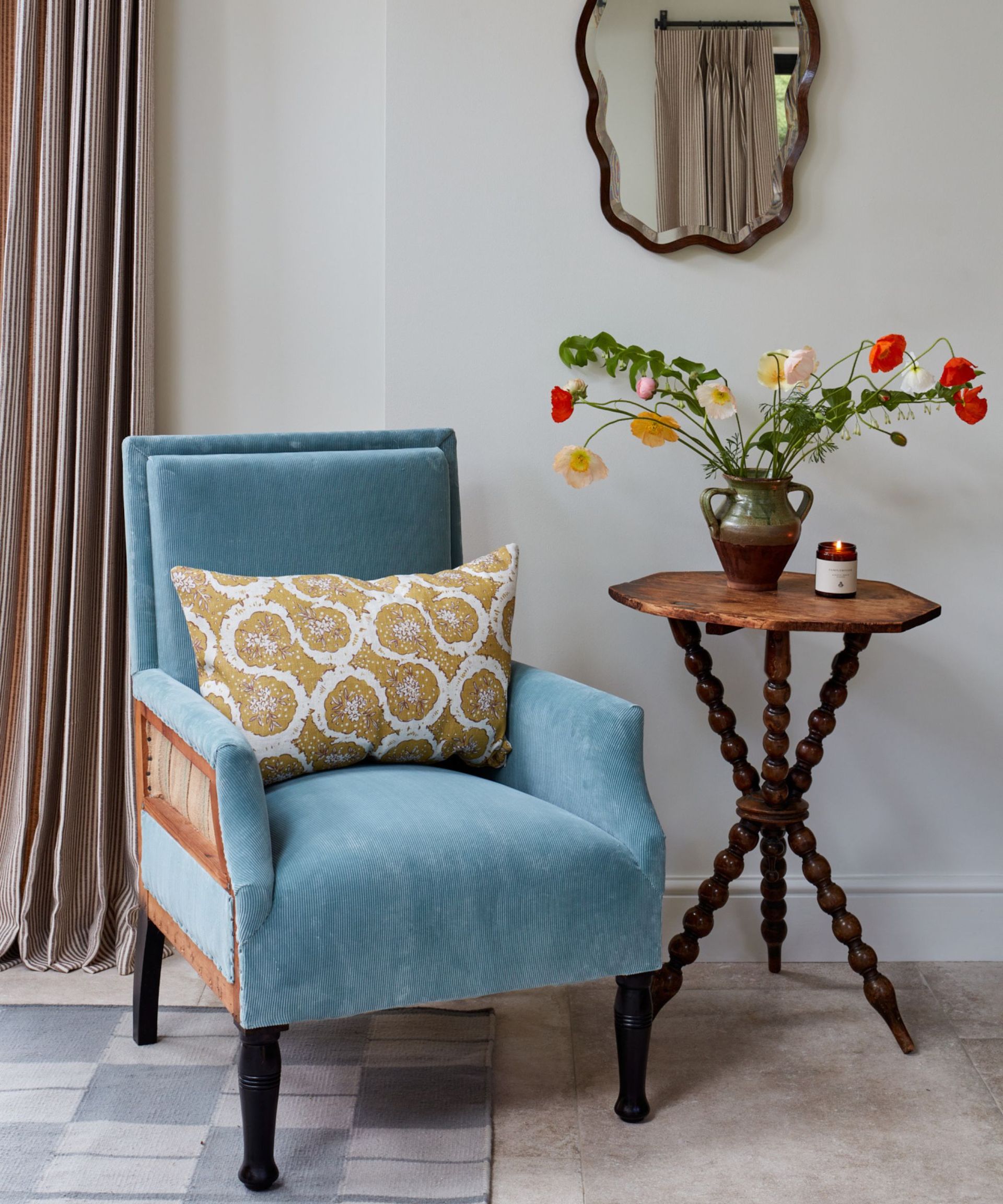 small vignette with a vintage style pale blue corduroy accent chair, an antique tripod bobbin side table with a vase with flowers and a wavy mirror hung above