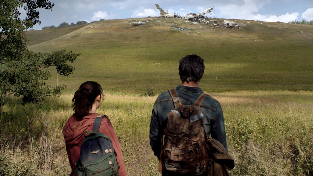 Ellie (Bella Ramsey) and Joel (Pedro Pascal) stand with their backs to the camera, looking at a hill in the distance, which has the wreckage of a crashed aeroplane on the top of it