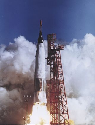 A rocket lifts off next to a red launch tower. White smoke fills the bottom two-thirds of the background before breaking to a blue sky. The rocket spits yellow-white fire from its engines as its chrome and frozen-white fuselage lift a black capsule skyward.