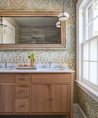 A bathroom with colorful floral wallpaper, a rectangular wooden mirror, a glass pendant light, a sink with wooden cabinets, and a window to the right