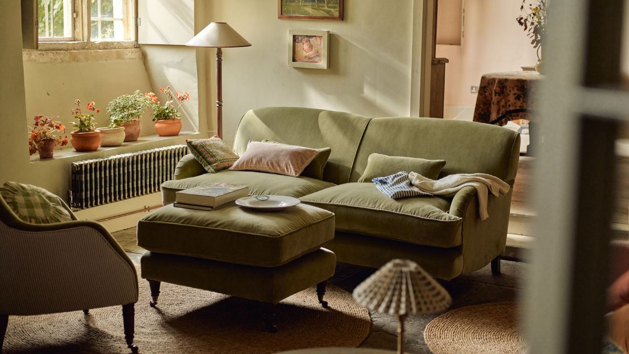 A sunlit living room with a green fabric sofa and matching footstool. 