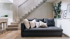 A bottom floor space with an open white staircase behind a blue sectional sofa. A small kitchen island off to the left hand side. 