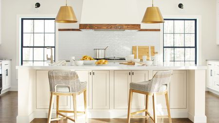 White kitchen with two large windows with black frames, a white subway tiled backsplash, an oven hood with wooden trim, a white kitchen island and walls painted in Benjamin Moore's Timid White.