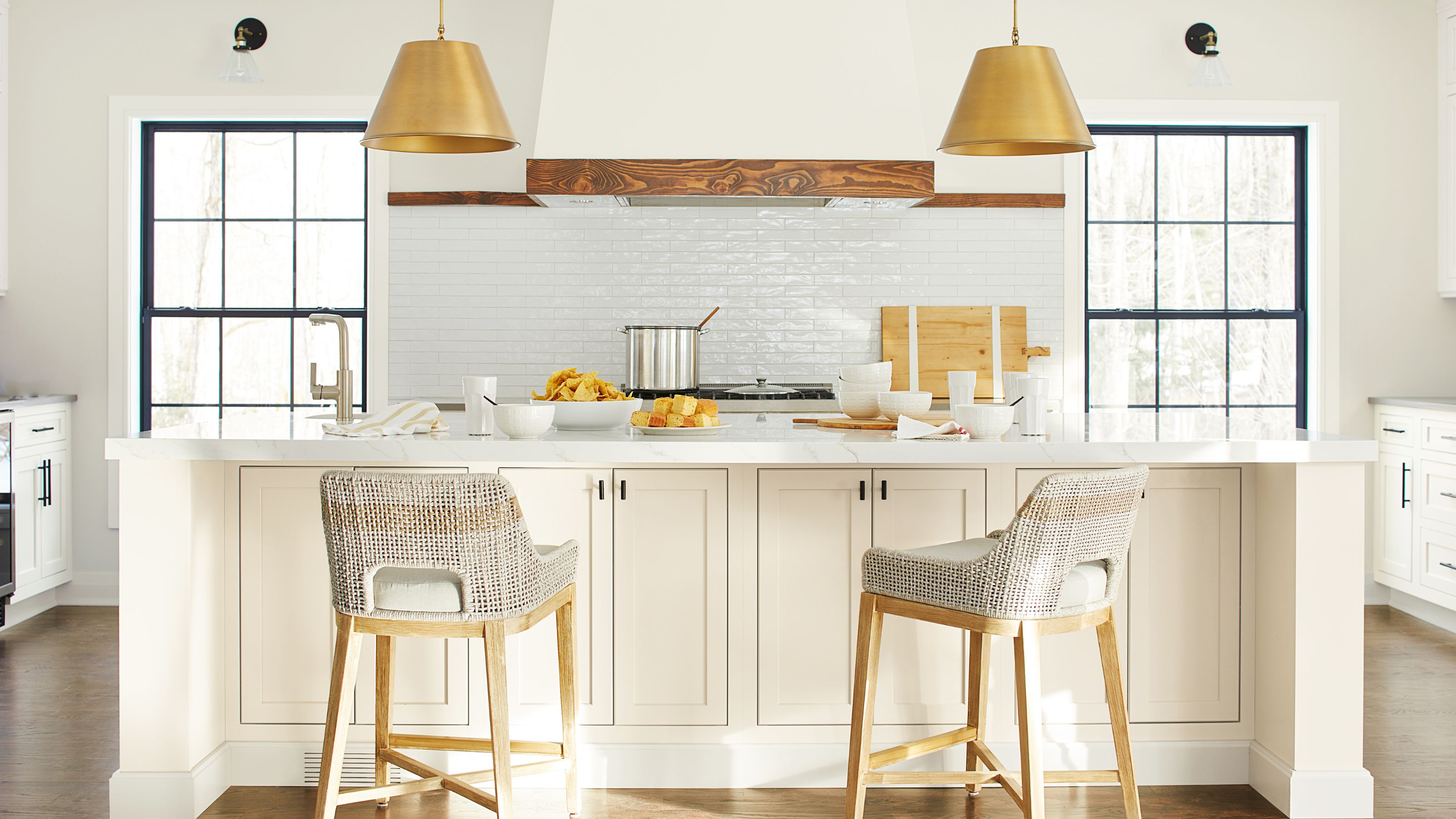White kitchen with two large windows with black frames, a white subway tiled backsplash, an oven hood with wooden trim, a white kitchen island and walls painted in Benjamin Moore's Timid White.