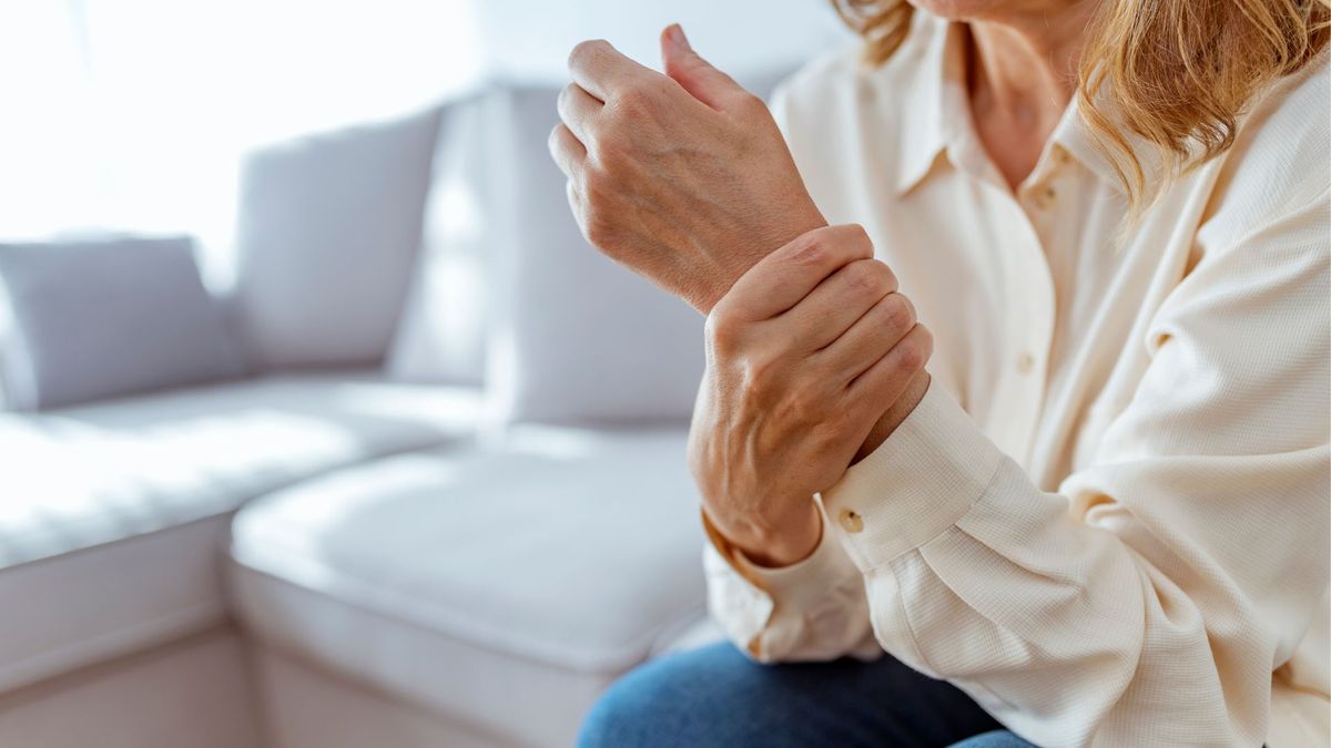 A close up of white woman&#039;s wrist, which she&#039;s holding as if experiencing pain or joint stiffness. She&#039;s sitting on a grey couch and wearing a white blouse