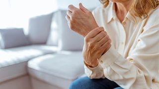 A close up of white woman's wrist, which she's holding as if experiencing pain or joint stiffness. She's sitting on a grey couch and wearing a white blouse
