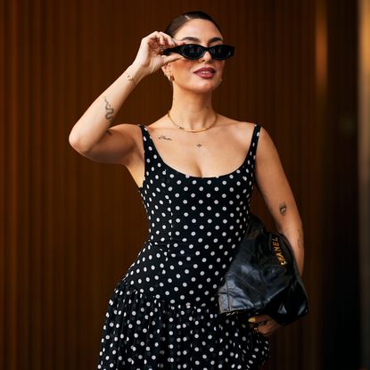 A woman at Paris Fashion Week wearing a black and white polka dot dress, black sunglasses, and black Chanel handbag.