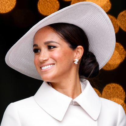 Meghan Markle wearing a white coat and matching hat with gold dots behind her