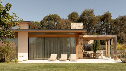A beautiful home with an outdoor patio featuring sun loungers and a dining space under a pergola by a green lawn