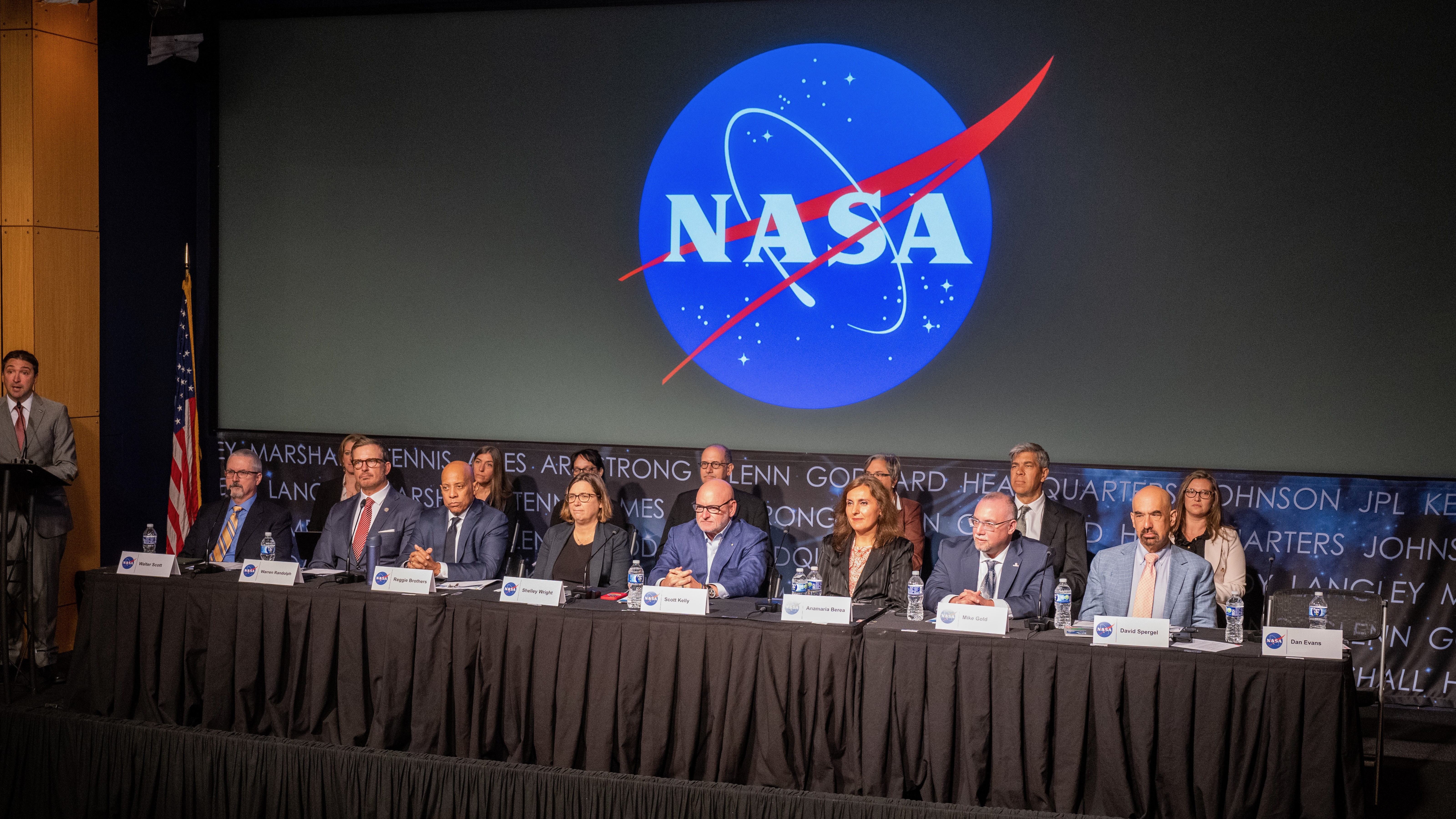 16 well-dressed people sit at a table in a conference room beneath a large nasa logo