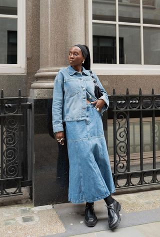 A denim skirt outfit is shown in a photo of a woman on the street wearing a black stretchy headband, silver earrings, a denim jacket, a black suede back, a denim skirt, and black loafers