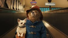 A still from the movie Paddington 2 of Paddington the bear wearing a blue coat and a red hat holding a little white dog on the escalator of the London underground.