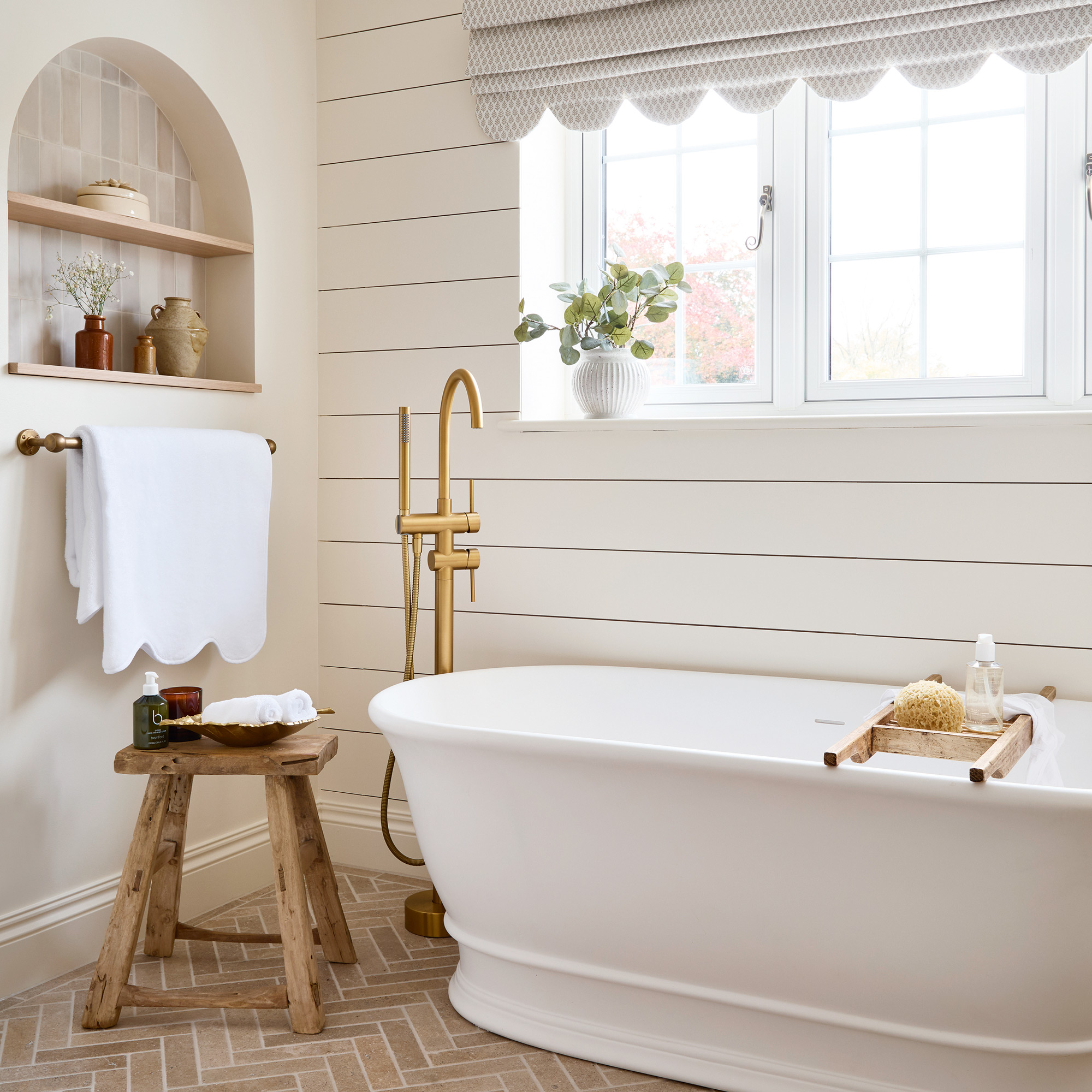 a neutral bathroom with horizontal wall panelling, a large white freestanding bath tub and brass fixtures, a scalloped edge roman blind and limestone herringbone floor tiles