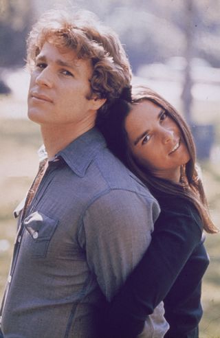 merican actors Ryan O'Neal and Ali MacGraw stand back to back outdoors in a still from the film, 'Love Story,' directed by Arthur Hiller, 1970.