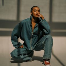 Maisie Richardson- Sellers crouched down wearing an oversize zip-up denim jacket and baggy jeans with red Adidas sneakers. She has her left hand raised to her mouth. 