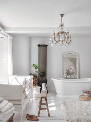 White bathroom with cozy farmhouse decor. There is a wood fireplace in the corner and a white love seat with a wooden bench in front