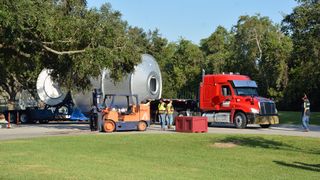 a red flatbed truck hauls a large silver cylinder on a tree-lined road