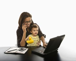 A woman sits at a computer with a toddler on her lap