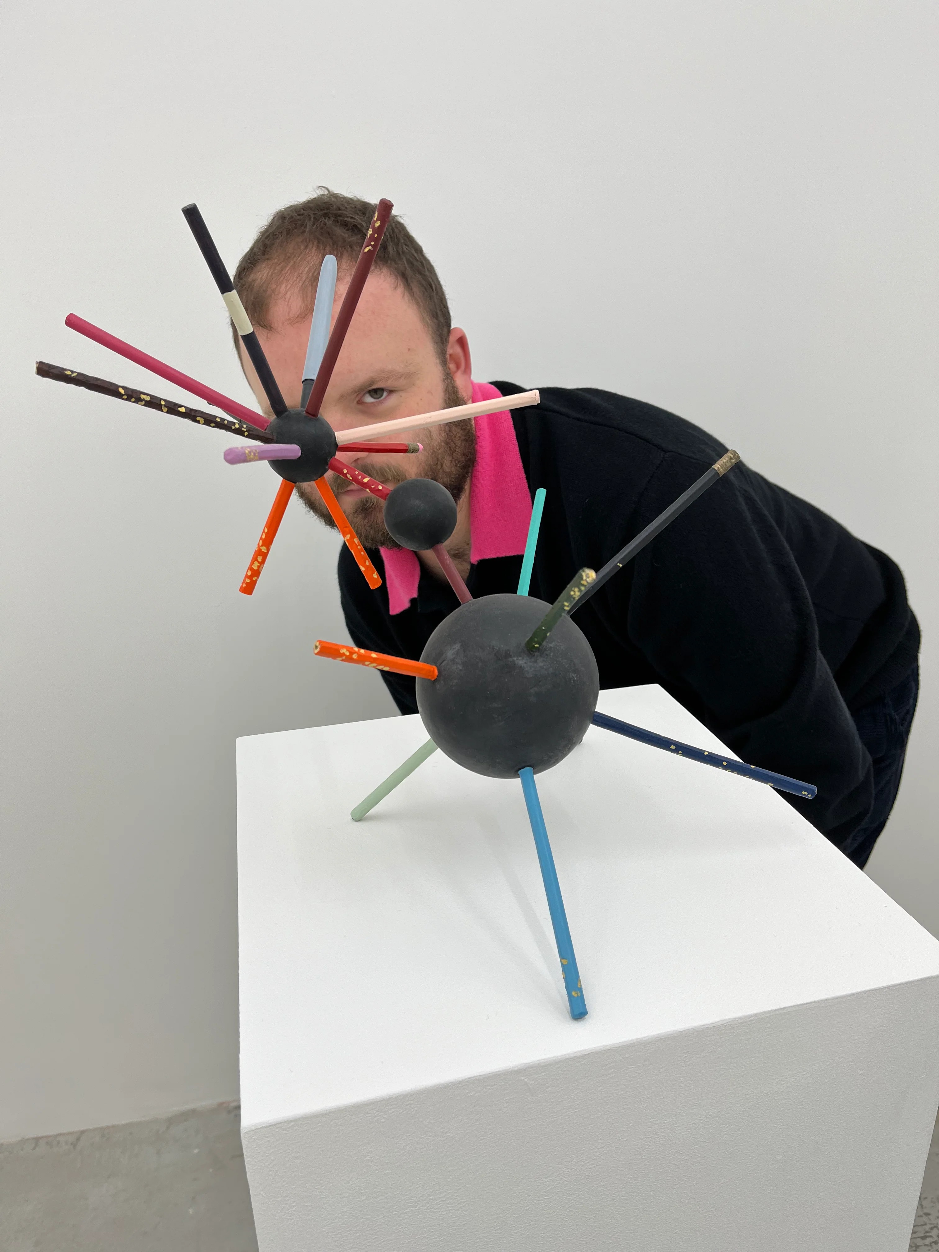 A man dressed in a pink polo shirt and a black jumper layered on top of it hides behind a foam and pencil sculpture in an industrial gallery space.