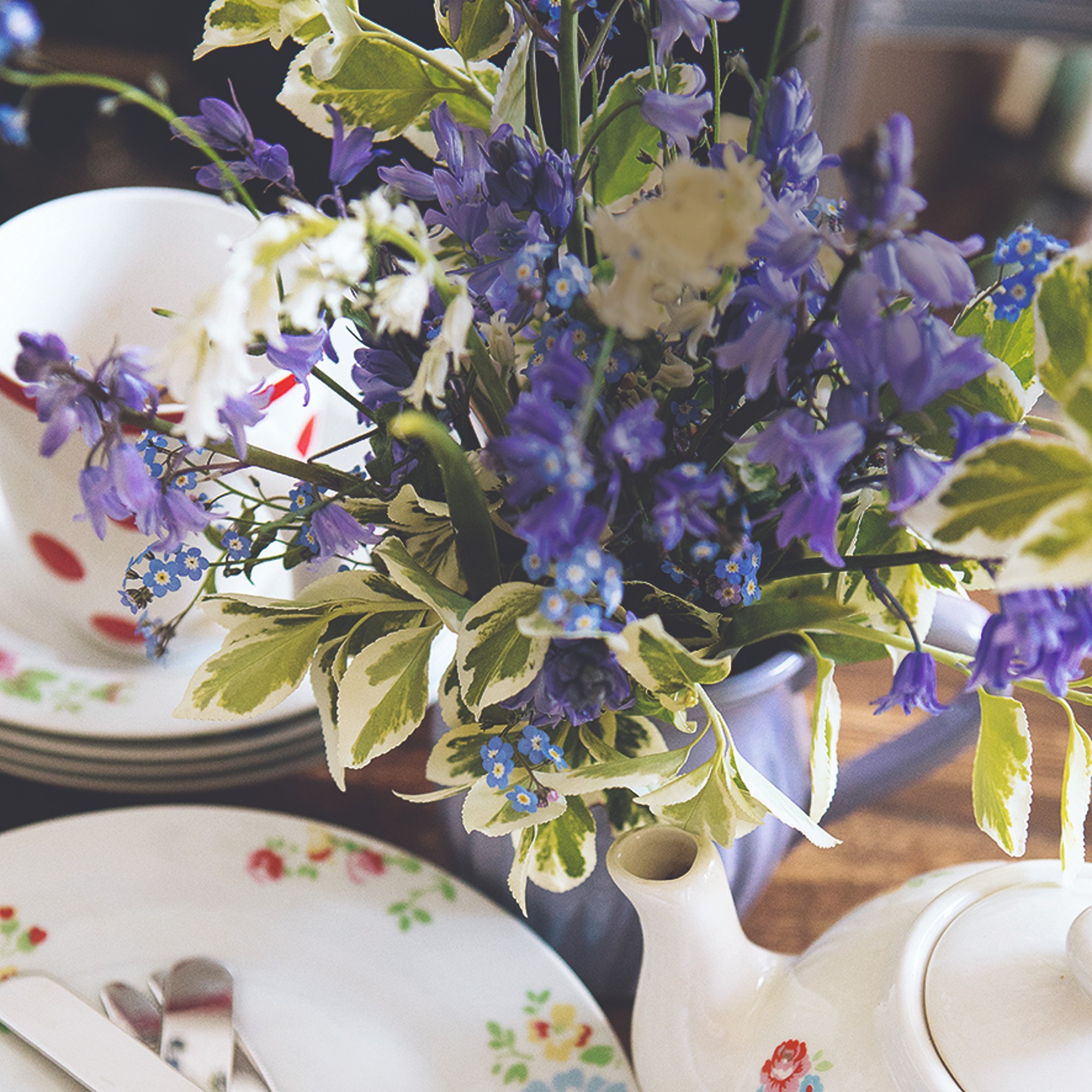 A vase of bluebells with a stack of plates, teacups and a teapot