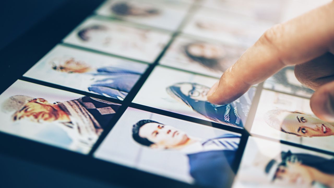 With only his finger showing, a man points at a photo of a woman among photos lined up on a tablet screen.