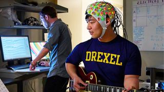 a man with tan skin and black hair wearing an EEG cap with colorful electrodes on it as he plays a guitar. A researcher stands in the background at a computer, watching brain waves on the screen