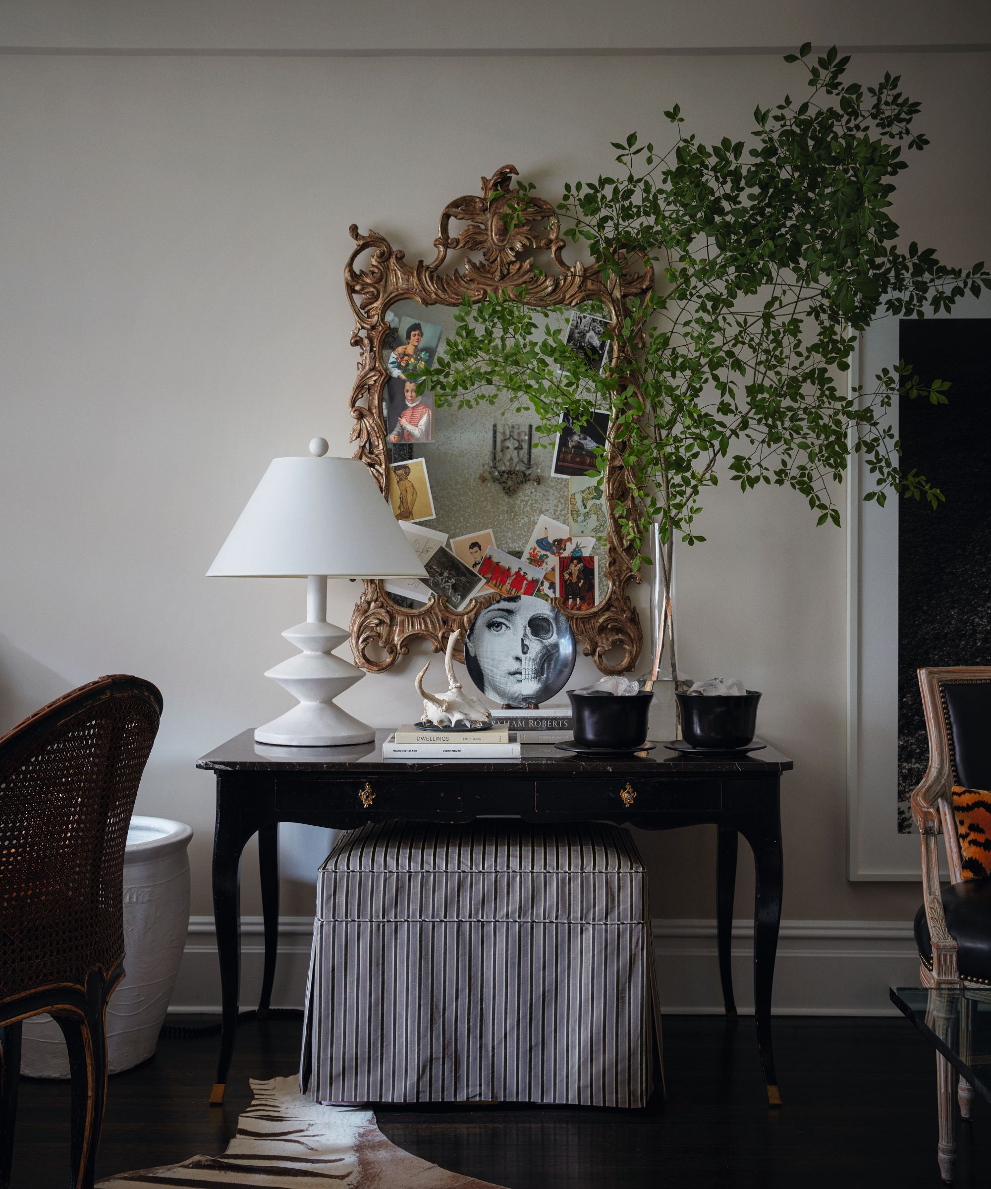 An ornate gold rococo mirror adorned with postcards hangs above a black console table, styled with a white geometric lamp, a decorative plate, and a tall leafy branch