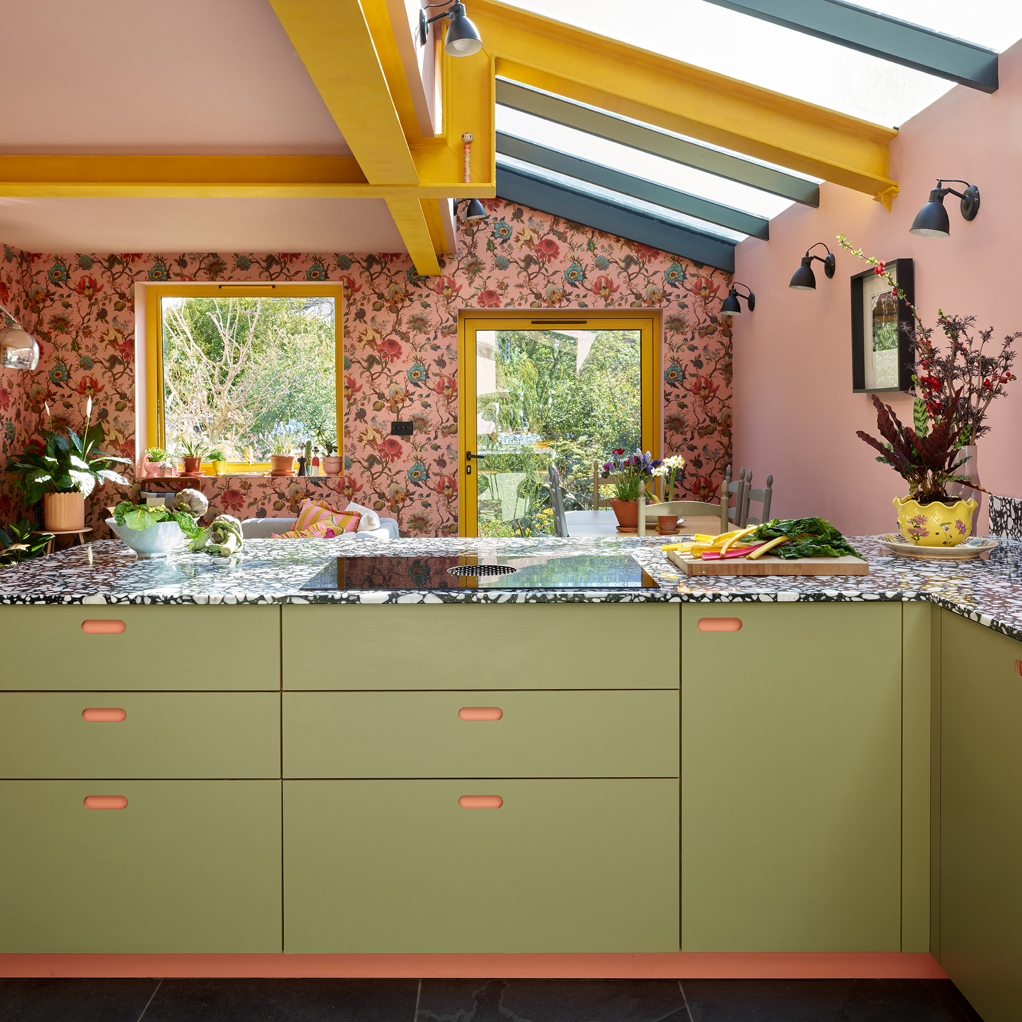 colourful kitchen with side extension pink ceiling yellow steel beams yellow framed window and glass door floral pink wallpaper and green kitchen cabinets with black and white worktop
