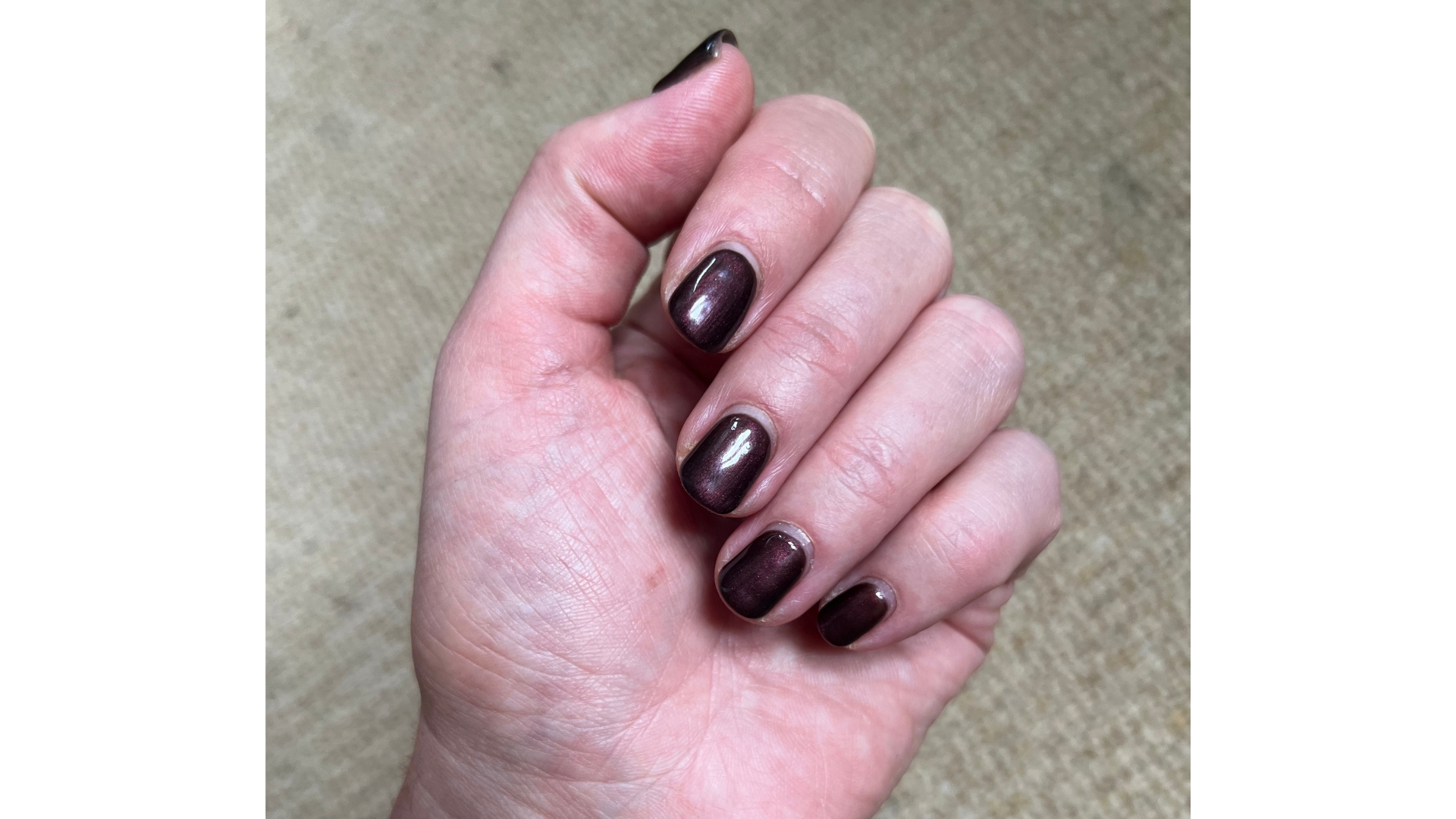 A close-up of Digital Beauty Editor, Fiona McKim&#039;s hand, showcasing her short and shimmery dark brown, cola-inspired manicure/ pictured in front of a neutral, textured carpet background