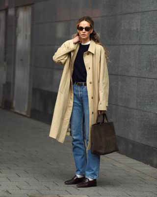 Mode-Influencerin Anouk Yve steht auf dem Bürgersteig und trägt eine ovale Sonnenbrille, einen Trenchcoat mit Kragen, einen schwarzen Pullover über einem weißen T-Shirt, eine braune Wildleder-Einkaufstasche, einen schwarzen Gürtel, Jeans mit geradem Bein, weiße Socken und Slipper