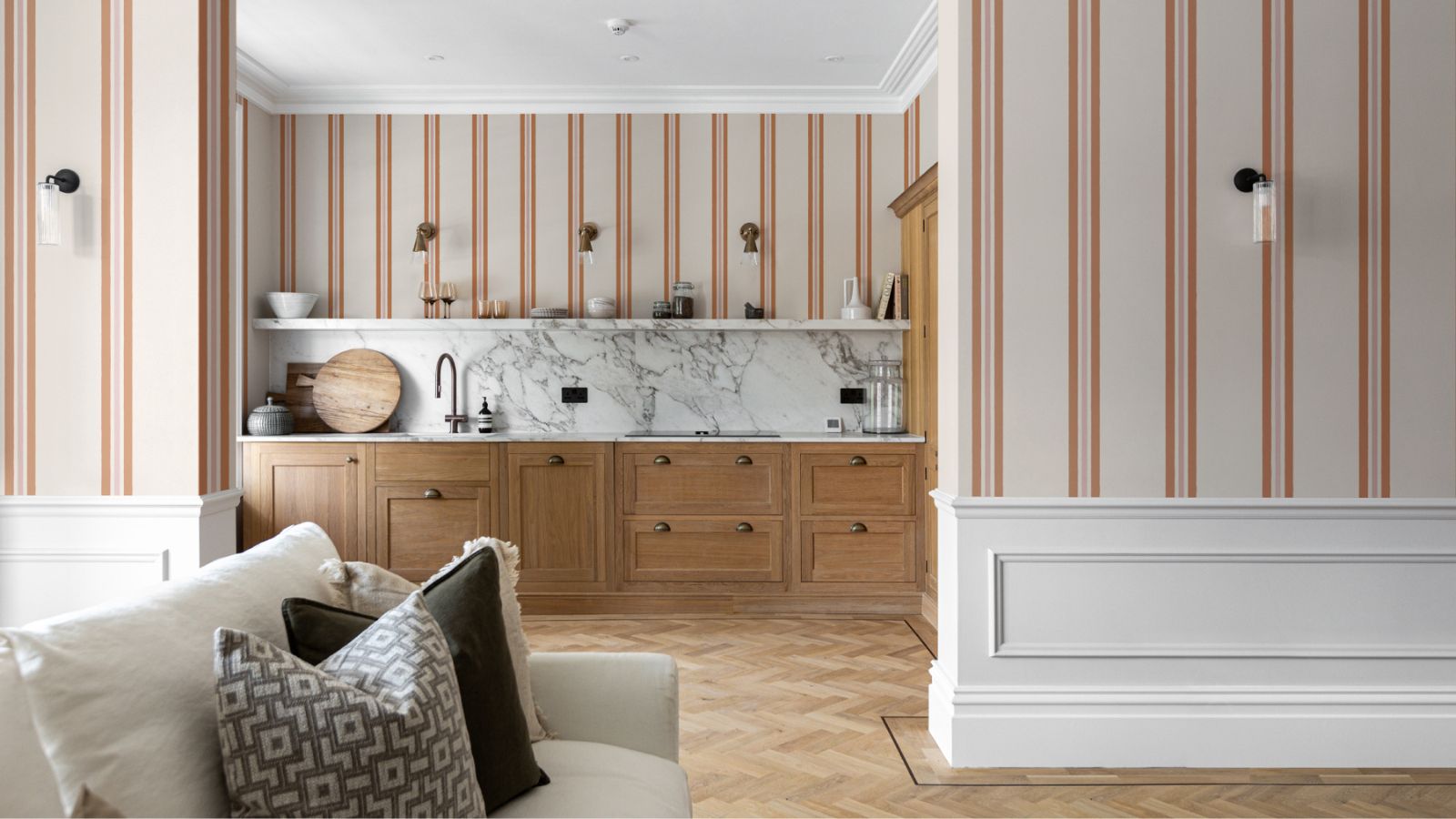 A stylish kitchen and living space featuring warm wooden cabinetry, marble splashback, and striped wallpaper in soft blush and terracotta. White panelling and herringbone flooring complete the elegant, timeless design.