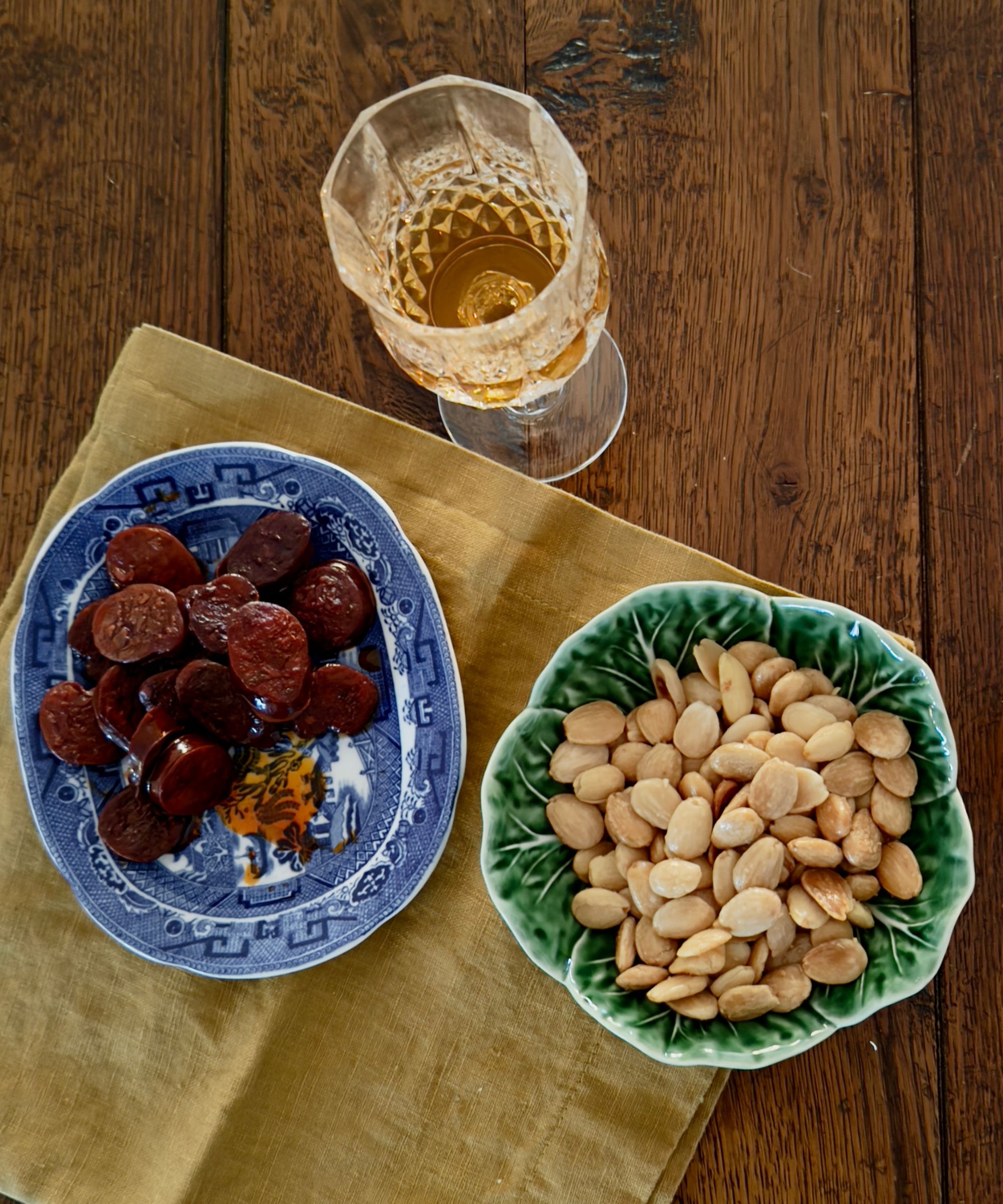 Bowl of sliced chorizo, a bowl of nuts and a glass of sherry on a wooden table with a napkin