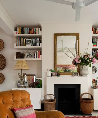 A white ceiling fan with white walls around it, colorful books on bookshelves, a fire place with flowers and wall art on top, and a tufted button orange seat with a throw pillow