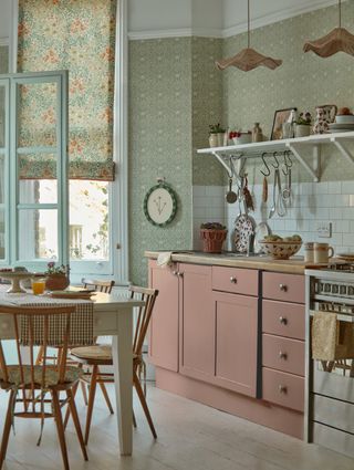 kitchen with green print wallpaper, floral blinds, blush pink cabinetry, open shelving, rattan pendants, kitchen table and chairs, range, white painted floorboards