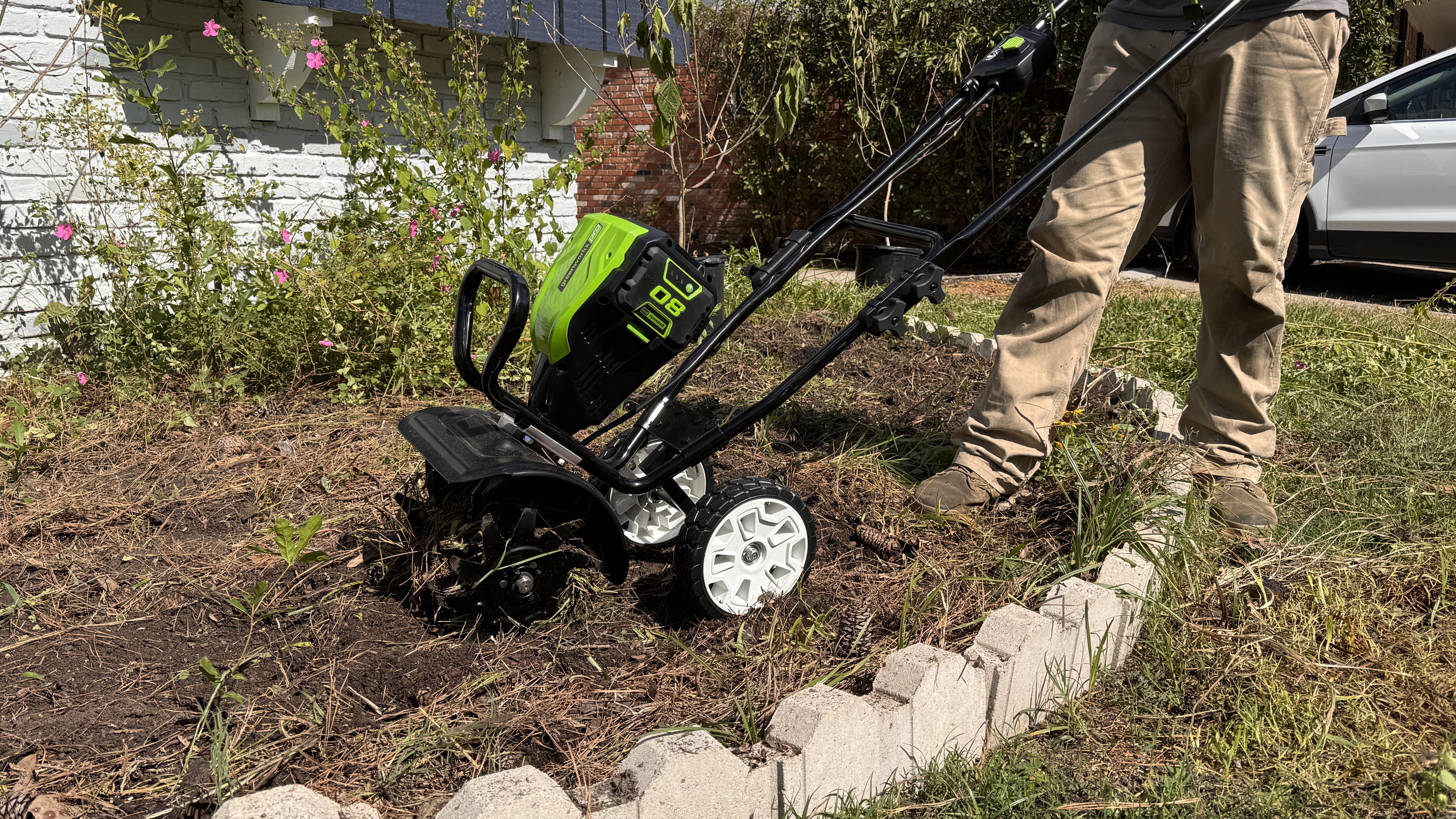 Man using GreenWorks tiller in garden bed with car in background