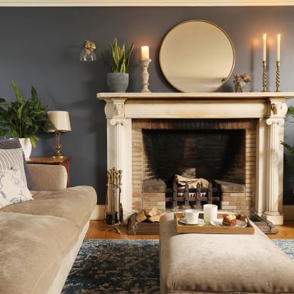 A dark-painted living room with a fireplace, lit candles and a beige velvet sofa