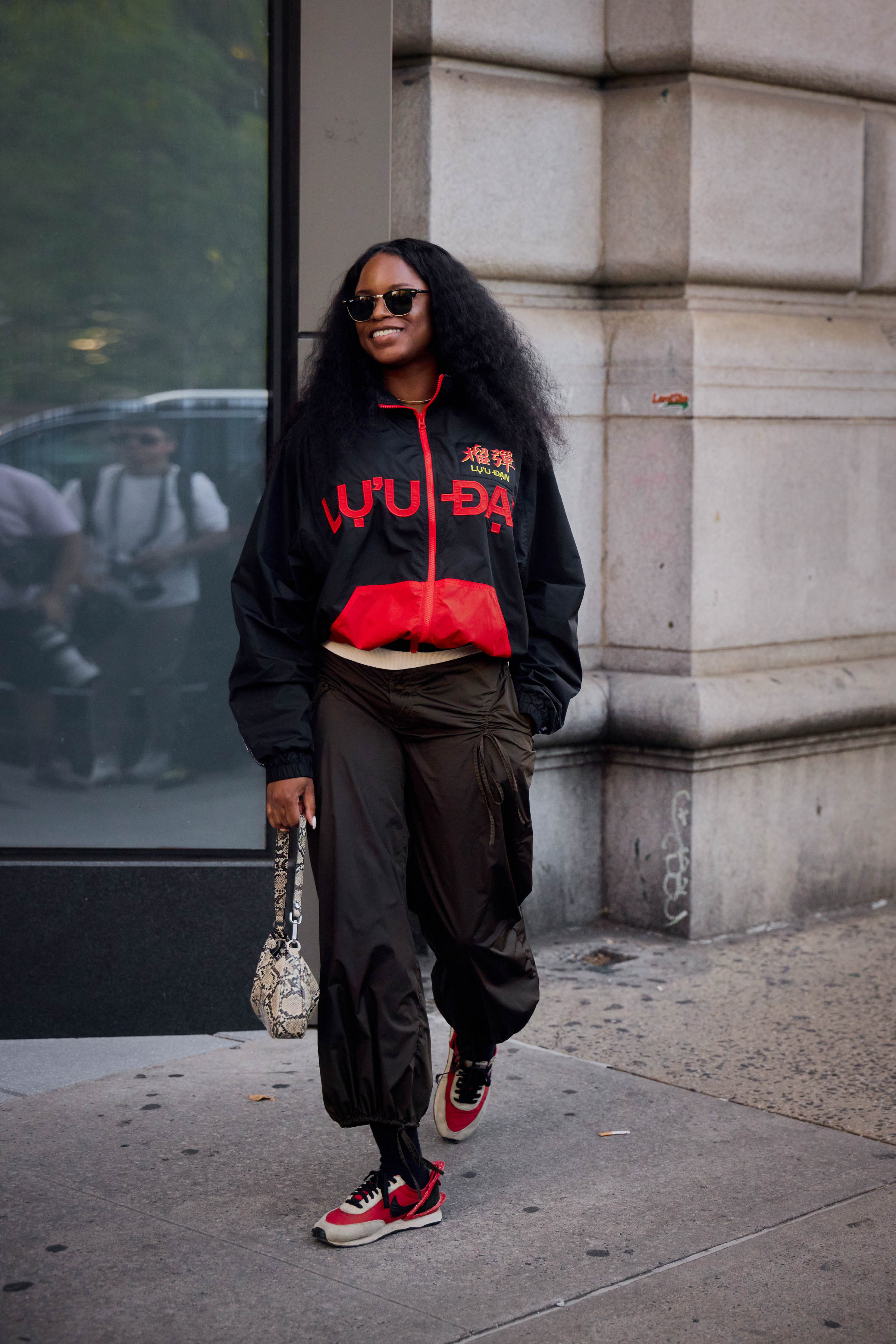 A woman wears red sneakers (one of the biggest sneaker color trends of 2026), black pants, a snakeskin bag, and a black sporty jacket.