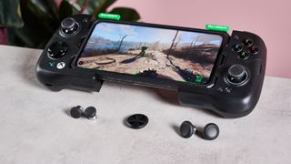 GameSir X4 Aileron on table with spare D-pad hat and analog stick caps, with plant and pink wall in background